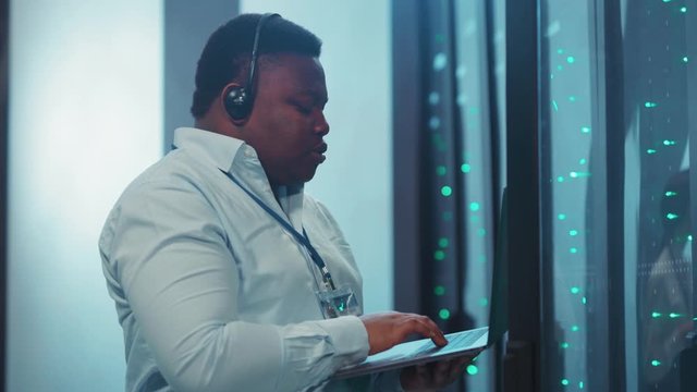 Busy african IT service support technician talking performing working tasks on notebook inspecting digital server rack cabinets in high tech computer security. Data center.