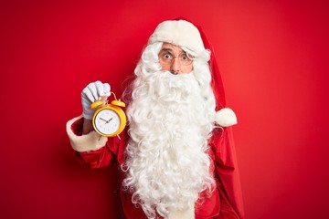 Senior man wearing Santa Claus costume holding alarm clock over isolated red background scared in shock with a surprise face, afraid and excited with fear expression
