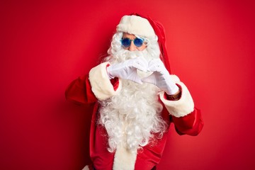 Middle age man wearing Santa Claus costume and sunglasses over isolated red background smiling in love showing heart symbol and shape with hands. Romantic concept.