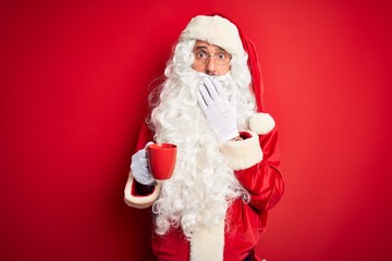 Senior man wearing Santa Claus costume holding cup of coffee over isolated red background cover mouth with hand shocked with shame for mistake, expression of fear, scared in silence, secret concept