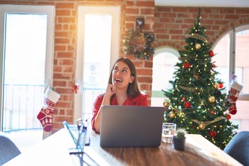 Beautiful woman sitting at the table working with laptop at home around christmas tree with hand on chin thinking about question, pensive expression. Smiling and thoughtful face. Doubt concept.