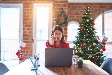 Beautiful woman sitting at the table working with laptop at home around christmas tree pointing displeased and frustrated to the camera, angry and furious with you