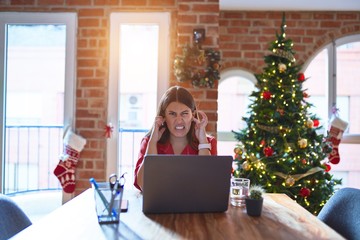 Beautiful woman sitting at the table working with laptop at home around christmas tree covering ears with fingers with annoyed expression for the noise of loud music. Deaf concept.