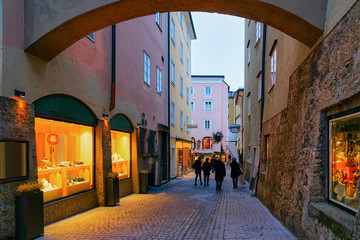 People at Narrow Street with shops and stores at Old city of Salzburg, Austria. Building architecture of Mozart town, Europe, winter. Panorama and landmark. Cityscape in Evening. Travel and tourism. - obrazy, fototapety, plakaty