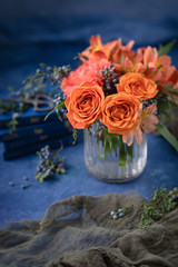 Vase of orange roses, carnations, and alstroemeria in clear vase on blue textured background; vintage scissors on small blue books in background; sprigs of blue cedar berries surround vase.