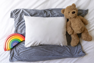 Blank white rectangle cushion laying on a grey scarf with a white duvet background and child's teddy and rainbow - nursery pillow mockup