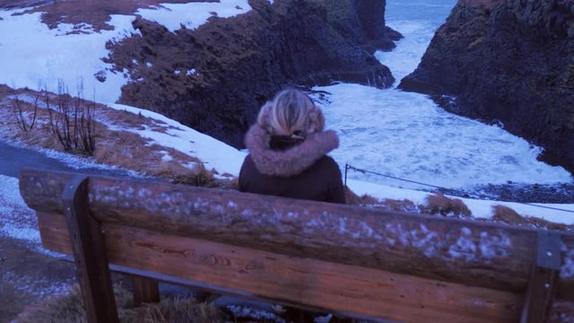 Iceland Female Sitting On Bench Looking At Cliffs And The Ocean In Arnarstapi In Winter 5