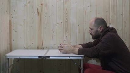 a man with a short haircut and beard drinks tea from a white mug, periodically puts the mug on a folding table, looks thoughtfully ahead, then gets up and leaves. red pants, Burgundy jacket with hood