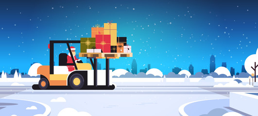 santa claus driving forklift truck loading colorful gift present boxes delivery and shipping concept merry christmas happy new year winter holidays celebration horizontal snowy sityscape background © mast3r