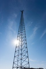 Bright sun shines behind radio tower antenna in a blue sky.