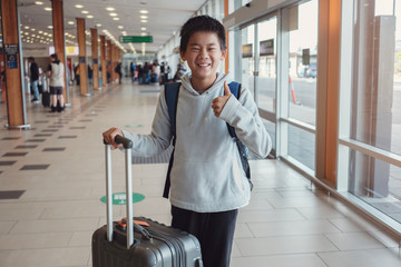 Happy mixed race Asian tween boy giving thumb up and holding his suitcase at the airport, preteen...