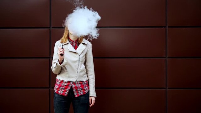 Vaping teenager. Young pretty white girl in casual clothes smoking an electronic cigarette opposite dark modern backgroun on the street in the autumn. Bad habit. Vape activity.