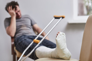 a man with a broken leg and crutches sits grabbing his head. Focus on the foot.