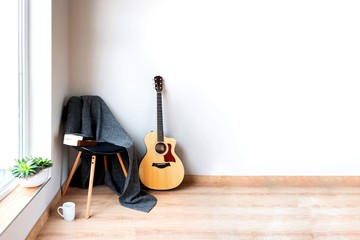 Contemporary home interior. Black chair covered with woolen gray blanket and acoustic guitar in front of an empty white wall. Time for hobby and leasure.