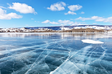 Sunny winter day on Lake Baikal. Blue clear ice with cracks in the Small Sea Strait. Wooden landmarks marked the ice road. On the shore of wooden houses of tourist camps