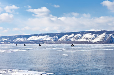 Sunny winter day on Baikal Lake. A group of tourists travels on snowmobiles on the ice of the frozen Small Sea Strait. Active winter holidays