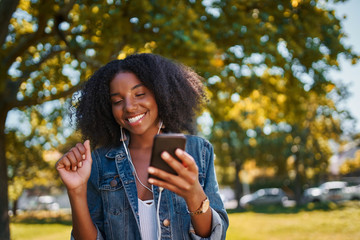 Portrait of an african american young woman enjoying the music on mobile phone through earphones in the park - Young black woman listening to music and dancing and having fun in the park outdoors