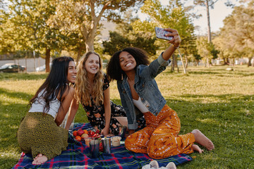 Group of happy diverse three female friends sitting together on blanket over the green grass enjoying at picnic taking selfie on smartphone in the park - obrazy, fototapety, plakaty