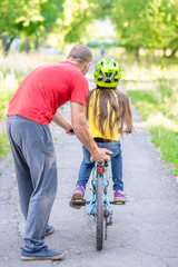Active family leisure. Father teaches his daughter to ride a bicycle in the summer park. Back view