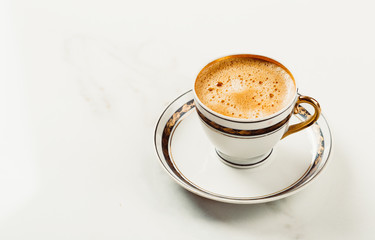 Cup of fresh americano or espresso coffee with golden foam froth on pile of brown raw coffee beans on white marble table background. Morning hot drink, coffee break, cope space