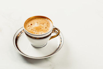 Cup of fresh americano or espresso coffee with golden foam froth on pile of brown raw coffee beans on white marble table background. Morning hot drink, coffee break, cope space