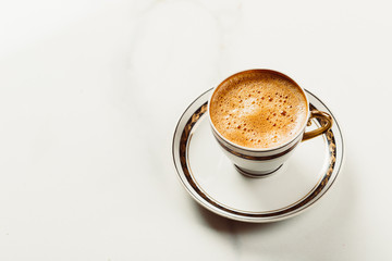 Cup of fresh americano or espresso coffee with golden foam froth on pile of brown raw coffee beans on white marble table background. Morning hot drink, coffee break, cope space