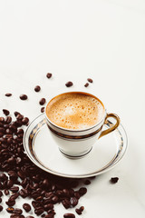 Cup of fresh americano or espresso coffee with golden foam froth on pile of brown raw coffee beans on white marble table background. Morning hot drink, coffee break, cope space