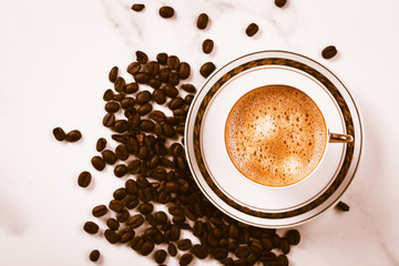 Cup of fresh americano or espresso coffee with golden foam froth on pile of brown raw coffee beans on white marble table background. Morning hot drink, coffee break, cope space