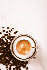 Cup of fresh americano or espresso coffee with golden foam froth on pile of brown raw coffee beans on white marble table background. Morning hot drink, coffee break, cope space