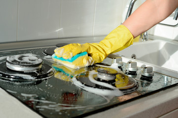 A hand in a yellow rubber glove washes a gas stove on a sunny day. Kitchen cleaning - obrazy, fototapety, plakaty