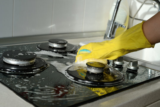 A hand in a yellow rubber glove washes a gas stove on a sunny day. Kitchen cleaning