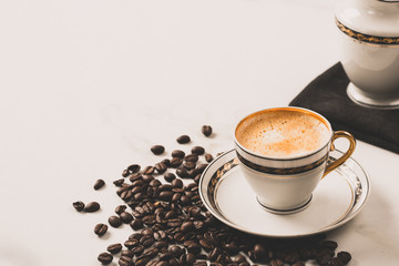 Cup of fresh americano or espresso coffee with golden foam froth on pile of brown raw coffee beans on white marble table background. Morning hot drink, coffee break, cope space