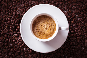 Cup of fresh americano or espresso coffee with golden foam froth on pile of brown raw coffee beans on white marble table background. Morning hot drink, coffee break, cope space