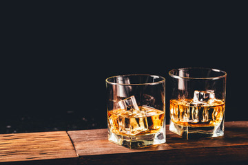 Two glasses of whiskey with ice cubes served on wooden board. Dark background, glossy golden color hard liquor on countertop with highlight. Night life bar relaxation concept