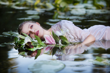 Beautiful sexy woman lying in the water in a white dress with a bouquet of lilies.