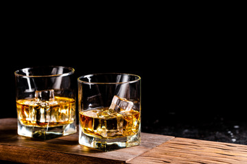 Two glasses of whiskey with ice cubes served on wooden board. Dark background, glossy golden color hard liquor on countertop with highlight. Night life bar relaxation concept
