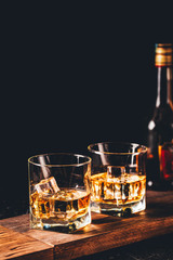 Two glasses of whiskey with ice cubes served on wooden board. Dark background, glossy golden color hard liquor on countertop with highlight. Night life bar relaxation concept