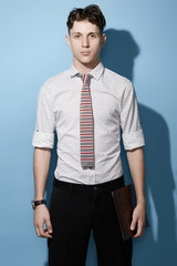 Fototapeta premium Medium full shot of a young dark haired man in a black trousers and white printed shirt with a knit tie with gray, red and white stripes. The man with a black watch is holding a leather folder. 