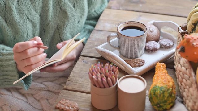 Girl drinks coffee and makes notes in a notebook. The atmosphere of comfort at home. A woman in a sweater has breakfast at the table.