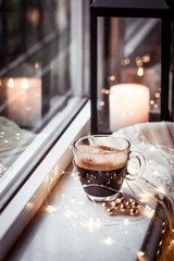 Glass cup of fresh strong americano coffee on marble windowsill in  winter morning with cinnamon stick, Christmas cozy garland bokeh twinkling lights, lantern and white cable knit scarf, copy space
