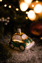 Christmas tree ornaments, tiny car on snowy table under the fir tree and bokeh lights with white candle in the background. Beautiful mysterious village decoration, greeting card, new year celebration 