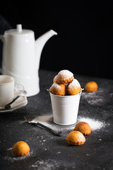 Fritter donuts balls or buns with cast sugar powder in white small bucket on dark background, delicious homemade easy dessert, breakfast or tea time concept