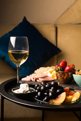 Cheese and fruits plate with glass of white wine on round tray, colorful and delicious charcuterie with snacks and wineglass for romantic evening, wine tasting, selective focus.