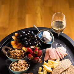 Cheese and fruits plate with glass of white wine on round tray, colorful and delicious charcuterie with snacks and wineglass for romantic evening, wine tasting, selective focus.