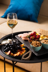 Cheese and fruits plate with glass of white wine on round tray, colorful and delicious charcuterie with snacks and wineglass for romantic evening, wine tasting, selective focus.