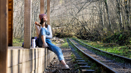 Young backpacker woman in denim overall is sitting on country train station alone and eating apple