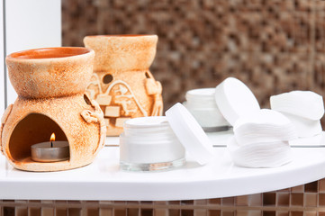 Brown aroma lamp with a burning candle, cotton pads and cream on a white shelf in the bathroom. Personal care for face and body skin. Mirror and shelf in the bathroom with jigsaw puzzle.