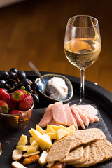 Cheese and fruits plate with glass of white wine on round tray, colorful and delicious charcuterie with snacks and wineglass for romantic evening, wine tasting, selective focus.
