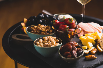 Cheese and fruits plate with glass of white wine on round tray, colorful and delicious charcuterie with snacks and wineglass for romantic evening, wine tasting, selective focus.