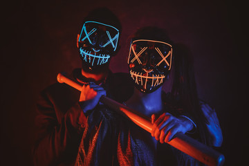 Man and woman in masks are posing for photographer with baseball bat in red and blue lights.